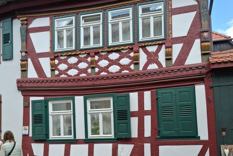Ein Fachwerkhaus mit rotbraunen Balken, von der Stra&szlig;e aus gesehen. Die Decke des Erdgeschosses ist durchgedr&uuml;ckt. Der erste Stock ist besonders sch&ouml;n gestaltet, mit gr&uuml;nen Fensterrahmen, Schnitzereien und Goldfarbe.