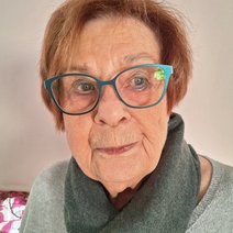 eine Seniorin, braune kurze Haare mit Scheitel rechts, mit runder, blauer Brille, die in die Kamera schaut