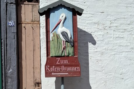 Ein Schild, mit der Aufschrift "Zum Roten Brunnen" und einem Pfeil nach links. Der obere Teil des Schildes ist ein Relief aus Holz, farbig bemalt. Es zeig einem Storch im Schilf. Dar&uuml;ber ist ein kleines Dach.