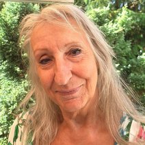 Eine lächelnde Frau, Mitte 60, mit langem blondem Haar, der Kopf ist nach links geneigt, sie steht vor grünen Büschen und Bäumen.