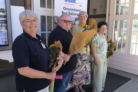 4 Menschen stehen vor der Trinkhalle und halten Blumen in der Hand.