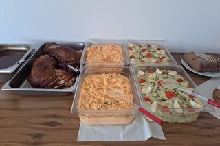 Verköstigung der Gäste mit Rollbraten und Salaten. Das Essen ist in flachen Gefäßen auf einem Tisch aufbereitet.