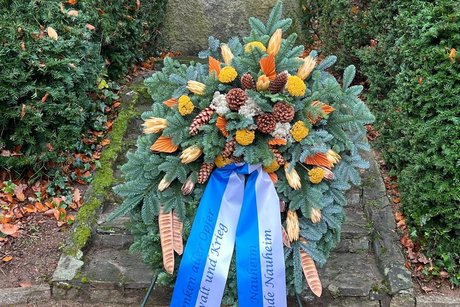 Auf dem Bild ist ein gro&szlig;er, handgebundener Gedenkkranz zu sehen, der auf einem Metallst&auml;nder steht. Er besteht aus frischen Tannenzweigen und ist mit Naturmaterialien wie Kiefernzapfen, getrockneten Bl&uuml;ten, gelblichen und orangefarbenen Zierpflanzen sowie dekorativen Holz- bzw. Schilfst&uuml;cken geschm&uuml;ckt. Der Kranz steht im Freien, vor einem Gedenkstein. Dieser ist von hohen, dunkelgr&uuml;nen Hecken eingerahmt. Die Inschrift des Steins lautet