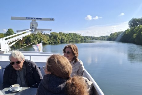 Ausblick vom Bug des Schiffes auf den Main. Blauer Himmel, ruhiges Wasser, B&auml;ume am Ufer rechts und links. Vorne links sitzen vier Fahrg&auml;ste am Tisch. Links oben ist ein Radarger&auml;t.