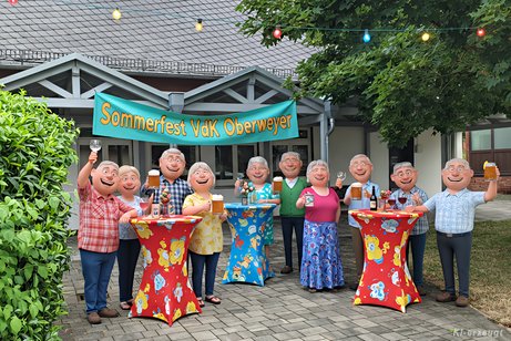 KI-erzeugtes Bild im Stil von Pixar-3D; 12 fr&ouml;hliche Menschen mit einem Getr&auml;nk in der Hand stehen an Stehtischen vor dem Pfarrheim.