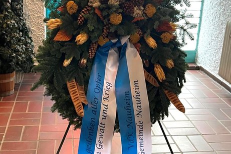 Wir gedenken aller Opfer von Gewalt und Krieg Auf dem Bild ist ein großer, festlich gebundener Gedenkkranz zu sehen, der auf einem Metallständer steht. Er besteht überwiegend aus dunkelgrünen Tannenzweigen und ist mit verschiedenen natürlichen Elementen dekoriert – darunter Kiefernzapfen, getrocknete Blüten, gelbliche und bräunliche Pflanzen sowie strukturierte Ziergräser.
