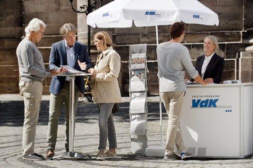 VdK-Infostand Ein VdK-Infostand in einer Fußgängerzone, eine Frau spricht mit einem Passanten, am Stehtisch daneben unterhalten sich Menschen.