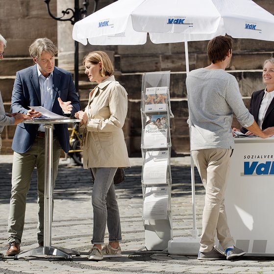 VdK-Infostand Ein VdK-Infostand in einer Fußgängerzone, eine Frau spricht mit einem Passanten, am Stehtisch daneben unterhalten sich Menschen.