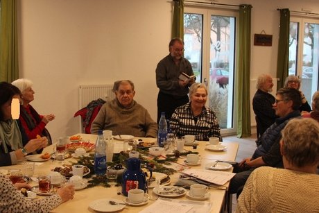 Um einen weihnachtlich gedekten Tisch, mit Kaffee und Gebäck, sitzen 7 Menschen. Dahinter steht Herr Hanke und lies aus einem kleinen Buch ein Gedicht vor.