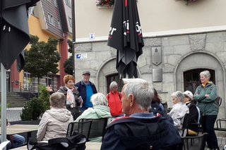 Die Besucher stehen und sitzen vor dem Rathaus und hören der Stadtführerin zu.