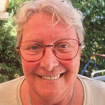 Eine lächelnde Frau, Mitte 60, mit kurzem, welligen grauen Haar, sie trägt eine große rote Brille und ein weißes Shirt, sie steht vor grünen Büschen und Bäumen.