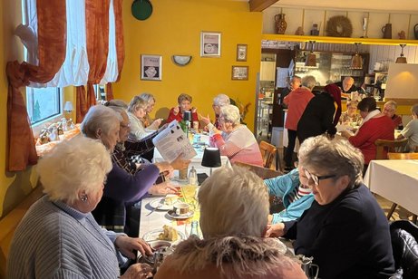 Frauenrunde beim Cafe Stelzer Das Bild zeigt eine gemütliche Zusammenkunft älterer Menschen in einem festlich dekorierten Raum, vermutlich eine Weihnachts- oder Adventsfeier. Was zu sehen ist: Viele Menschen sitzen an mehreren Tischen, trinken Kaffee oder Tee und unterhalten sich. Die Stimmung wirkt warm und gesellig. In der Raummitte stehen zwei Personen, die jeweils Körbe mit kleinen Geschenken oder dekorierten Flaschen halten – möglicherweise werden gerade Präsente verteilt. Rechts steht ein großer Tisch voller weihnachtlicher Dekorationen, darunter kleine Weihnachtsmänner, Kerzen und festliche Arrangements. Die Wände sind gemütlich dekoriert, u. a. mit Bildern, alten Küchenutensilien und einem Wandregal. Die Beleuchtung ist warm, was zur weihnachtlichen Atmosphäre beiträgt. Insgesamt wirkt es wie eine liebevoll organisierte Adventsfeier in einem Gemeinschaftsraum oder Vereinslokal.
