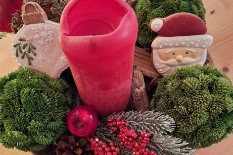 Ein weihnachtsgesteck mit roter Kerze, Tanne, Keks-Weihnachtsmanngesicht, Keks-Tannenbaumkugel in weiß und grüne Pflanzenkugeln