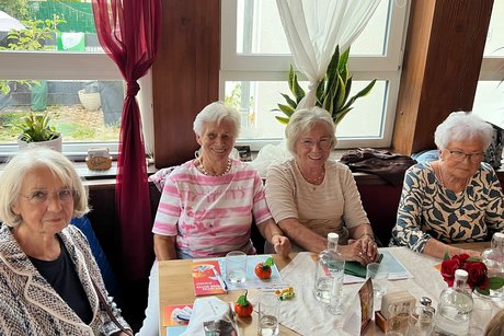 Das Bild zeigt eine Gruppe von vier Personen, die an einem festlich gedeckten Tisch in einem hellen Gastraum oder Restaurant sitzen.