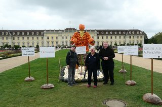 3 Vorstandsmitglieder stehen vor der ersten Kürbisfigur, umgeben von Schildern. Im Hintergrund ist das Schloss.