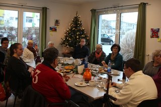 Um einen festlich gedeckten Tisch sitzen 11 Personen. Dahinter, in einer Ecke zwischen zwei Fenstern, steht ein Weihnachtsbaum.