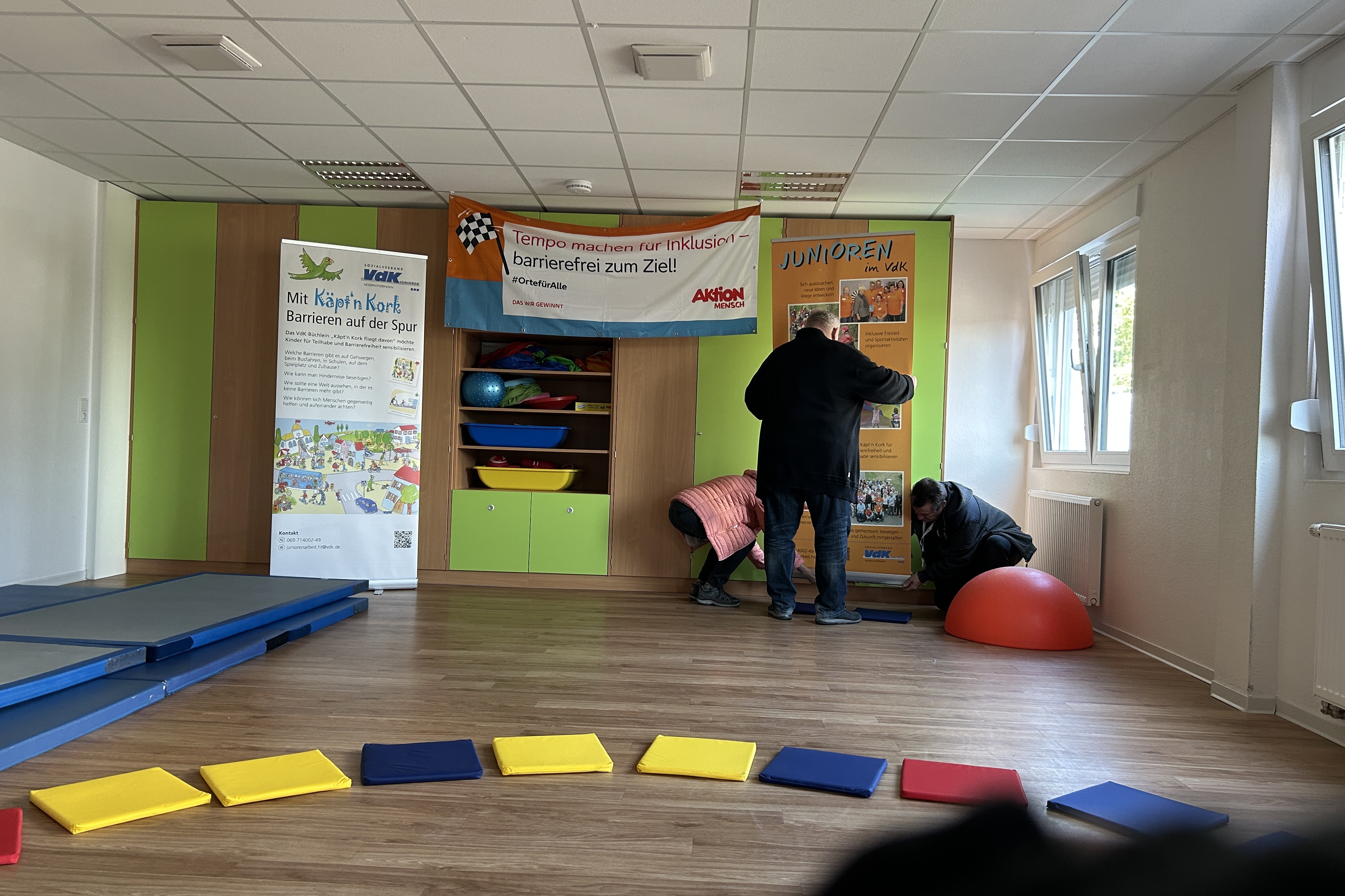 Das Bild zeigt die Vorbereitung f&uuml;r den Parcours, einen gro&szlig;en Raum vorne mit Sitzkissen im Halbkreis, hinten einen Mann der Rollups aufbaut