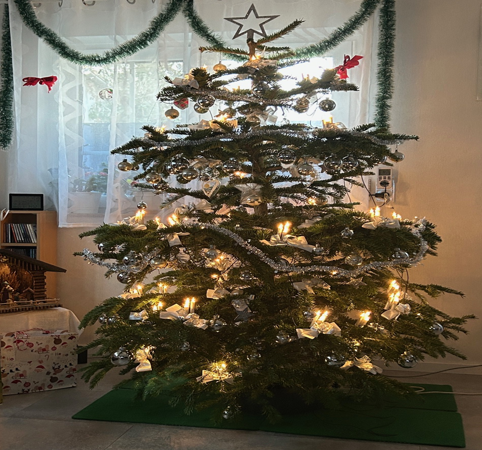 Das Bild zeigt einen Geschmückten Weihnachtsbaum mit Silbernen Weihnachtskugeln, leuchtende Kerzen und weiterem Baumschmuck